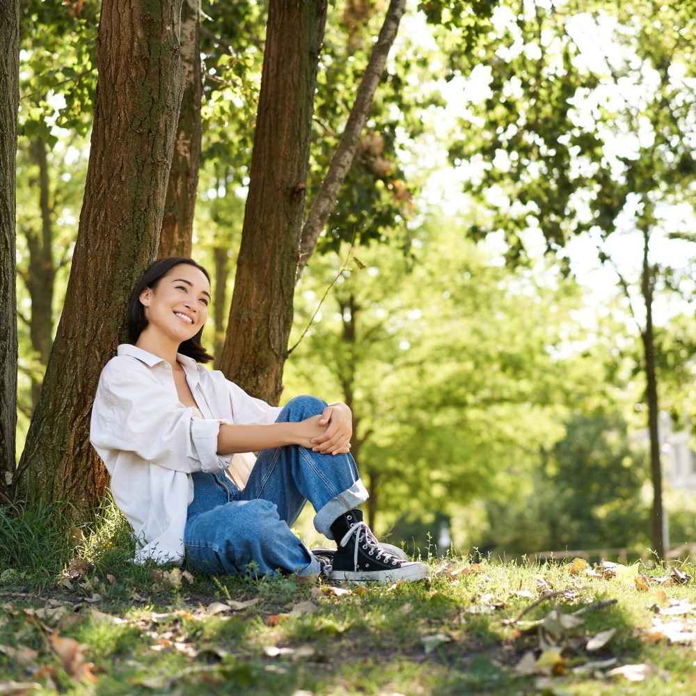 Beautiful smiling girl sits near tree in park, enjoying nature outdoors, relaxing and resting on fresh air. Copy space