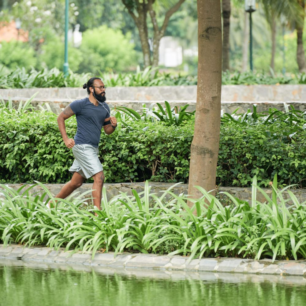 Bearded Indian sportsman in glasses and activewear running outdoors and listening to music with earphones