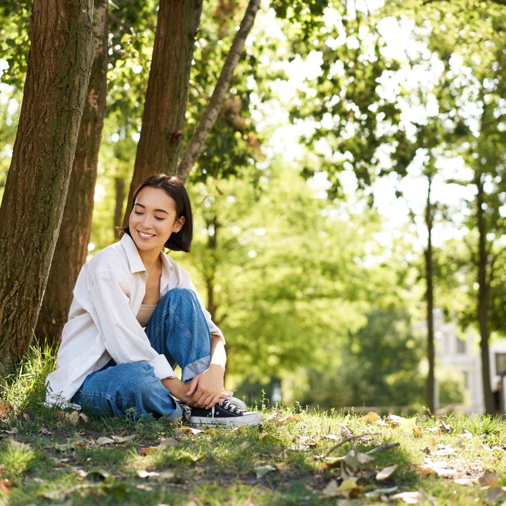 Portrait of beautiful asian woman resting near tree, relaxing in park, smiling and looking happy. Copy space