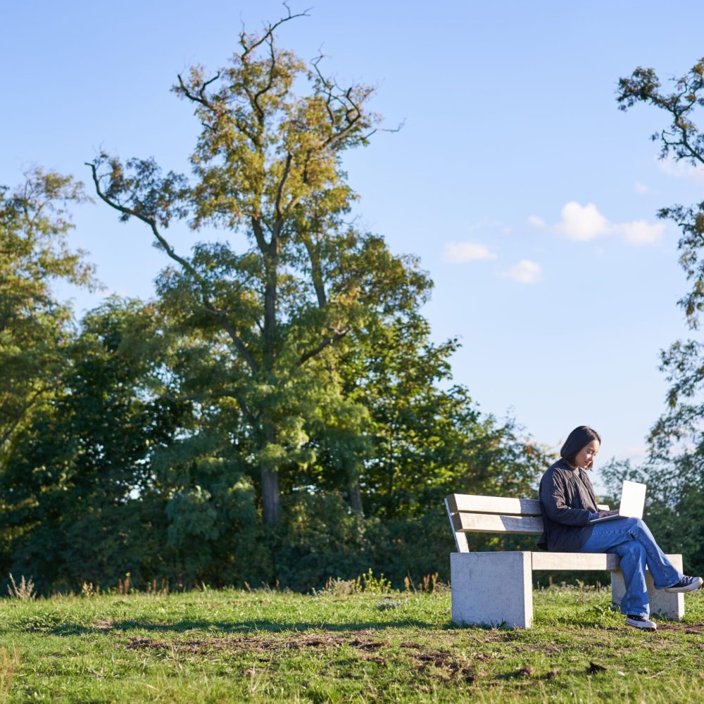Side view of young woman sitting alone in park on bench, using her laptop to study or work remotely from outdoors.